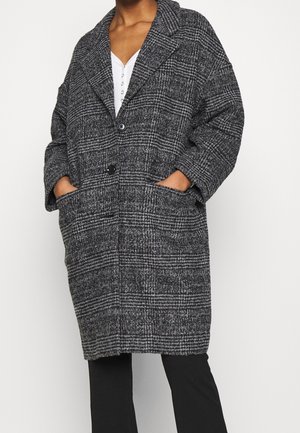 Personne portant un long manteau en laine à carreaux gris et noir avec de grands revers, deux boutons, poches avant, sur un pantalon noir et un haut blanc.