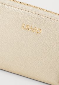 Busta in pelle beige dalla texture con cerniera dorata e logo impresso "LIU•JO" sul davanti. Design compatto con una finitura morbida.