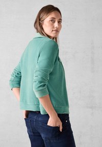 Turkos sweatshirt med böjd nederkant, ribbade manschetter och en mjuk textur, i kombination med mörkblå jeans. Modellen är placerad från sidan.