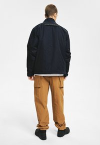 Granatowa kurtka z wysokim kołnierzem, zestawiona z jasnobrązowymi spodniami typu cargo. Kurtka ma etykietę z tyłu i luźny krój.