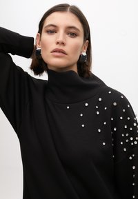 Schwarzer Sweatshirt mit hohem Kragen, verziert mit silbernen Strasssteinen auf einer Schulter. Glatter Stoff mit lockerer Passform.