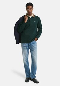 Polo a maniche lunghe verde scuro con colletto beige, abbinato a jeans azzurri e scarpe nere, che mostrano un look classico e informale.