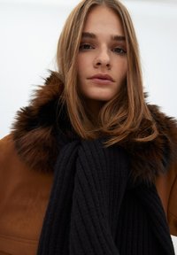 Brun mockaskinnjacka med pälskrage, tillsammans med en tjock svart ribbad scarf. Mjuka texturer och rika färger framhäver ensemblen.