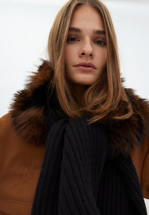 Chaqueta de ante marrón con cuello de piel, combinada con una gruesa bufanda negra de canalé. Las texturas suaves y los colores ricos destacan el conjunto.