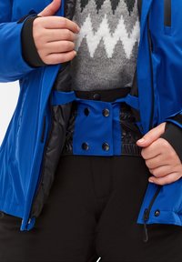 Chaqueta de invierno azul con acentos negros, que presenta un zipper, cierre a presión y cintura elástica. Lleva encima un suéter gris con estampado.