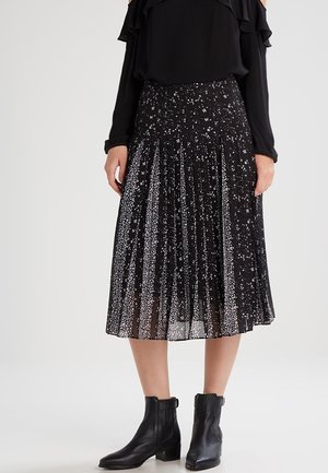 Femme portant une jupe midi plissée noire avec des taches blanches, un haut noir à manches longues avec des volants et des bottines noires, debout sur un fond blanc.