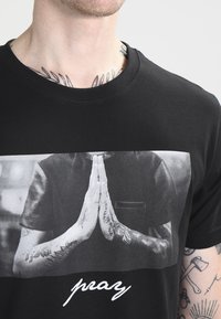 Camiseta de algodón negra con un gran gráfico gris de manos en posición de oración, con el texto cursivo blanco "reza" debajo.