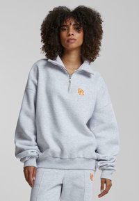 Grå fleece-pull med halv glidelås og ribbestrikkede mansjetter. Har en oransje logo på brystet og en avslappet passform. Myk tekstur.