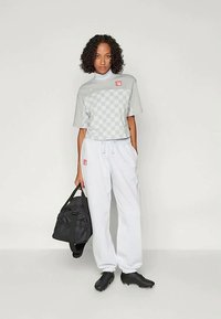 Kvinde med krøllet hår iført gråternet kortærmet top, hvide sweats, sorte sportssko, som holder en sort duffeltaske mod hvid baggrund.