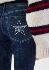 Jeans de ganga azul escuro com um bordado branco em forma de estrela no bolso de trás, apresentando o texto 'tommy girl', e com punhos de suéter de malha texturizada.