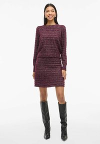 Maglione bordeaux a maglia con motivo testurizzato abbinato a una gonna aderente. La modella indossa stivali neri al ginocchio, presentando un look elegante e moderno.
