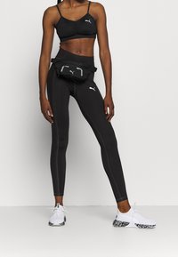 Czarny top z krótkim rękawem i legginsy z wysokim stanem o dopasowanym kroju, w połączeniu z czarną saszetką na biodra i białymi trampkami z wzorzystą podeszwą.