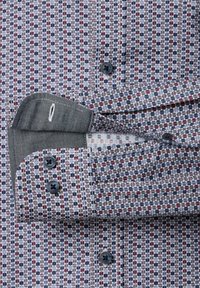 Langarmshirt met geometrisch hexagoonpatroon in rood, blauw en wit, voorzien van donkere knopen en een gedeeltelijk losgeknoopte manchette.