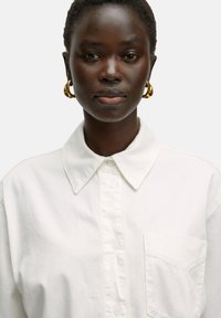 Witte blouse met kraag, borstzak en een zachte katoen structuur. Gekleed met grote gouden oorringen.