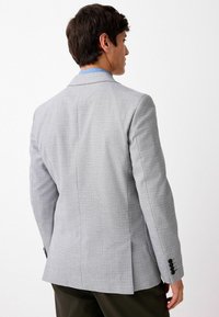 Grau strukturierter Blazer mit konturierten Revers, einem Schlitz auf der Rückseite und schwarzen Knöpfen an den Ärmeln. Lässiger Kragen darunter.