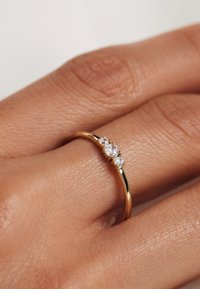 Gold ring with a smooth band featuring three round, clear gemstones set in prongs. Textured skin of a hand visible in background.