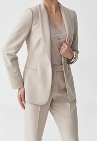 Costume beige clair comprenant un blazer ajusté à col cranté, un pantalon droit assorti et un haut simple à col en V en dessous.
