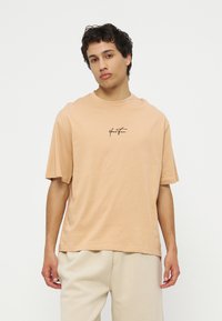 Νέος άνδρας με κοντά κατσαρά μαλλιά που φοράει μπεζ φαρδιά μπλούζα με το κείμενο "Your Turn" και ανοιχτόχρωμο παντελόνι, στέκεται μπροστά σε λευκό φόντο.