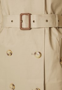 Beżowy płaszcz trencz z paskiem w talii, ozdobiony dużymi, okrągłymi, żółtymi guzikami. Gładka faktura tkaniny i dwurzędowy fason.