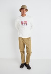 Chemise blanche à manches longues avec un imprimé graphique coloré au centre, associée à un pantalon marron clair et des chaussures marron, le tout agrémenté d'un chapeau bucket beige.