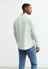Camisa de rayas verde claro, de mangas largas, con cuello, puños abotonados, hecha de tela suave, combinada con jeans azules. Sin patrones visibles en los jeans.