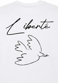 Witte t-shirt met een zwarte omtrektekening van een duif die een olijf tak vasthoudt, met het woord "Liberté" in een elegante schrift erboven.