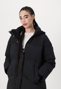 Black padded coat featuring a high collar, zip and snap closure, two side pockets, and a textured fabric. The design is oversized with a hood.