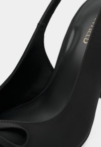 Close-up of a black high-heeled shoe with a bow detail near the toe and a slingback strap.