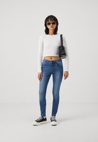 Top corto de manga larga blanco, jeans ajustados azules, bolso negro, zapatillas negras y blancas, combinados con gafas de sol azules de gran tamaño.