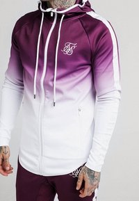 Mies pukeutunut liilasta valkoiseen liukuvärjättyyn vetoketjulliseen huppariin, jossa valkoiset kiristysnyörit, ja hänellä on yllään vastaavat liilat housut, joissa valkoiset sivuraidat.