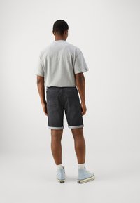 Man som bär lätt grå t-shirt, mörka uppvikta denimshorts, vita strumpor och ljusblå hög-topp sneakers, stående med ryggen vänd.