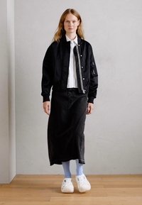 Veste bomber noire portée par-dessus une chemise blanche à col, accompagnée d'une longue jupe noire et de chaussettes bleu clair, le tout terminé par des sabots blancs.