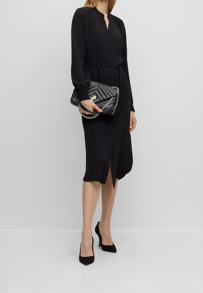 Femme portant une robe midi noire à manches longues et des talons noirs, tenant un sac à main noir matelassé avec une bandoulière chaîne argentée.