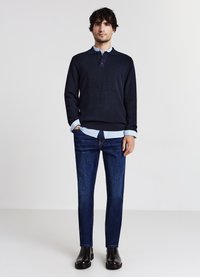 Maglione a maglia blu navy con colletto e tre bottoni, indossato sopra una camicia azzurro chiaro, abbinato a jeans blu scuro e stivaletti neri.