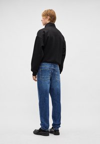 Sudadera negra de gran tamaño, vaqueros azules de cintura alta y zapatos negros gruesos, vistos desde atrás sobre un fondo liso.