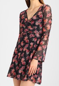 Schwarzblumiges Kleid mit V-Ausschnitt, transparenten langen Ärmeln, pinken und roten Rosenmustern sowie einem ausgestellten Rock. Leichte, fließende Stoffqualität.