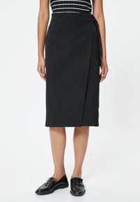 Zwarte wikkelrok van gladde stof, op knielengte, met een zijstrikdetail. Gecombineerd met zwarte instappers voor een complete look.