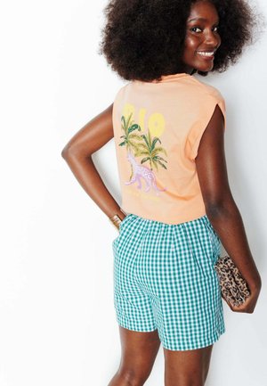 Femme aux cheveux bouclés souriant par-dessus l'épaule, portant un top sans manches couleur pêche avec imprimé léopard et palmier, short vert à carreaux, tenant une pochette imprimée léopard.