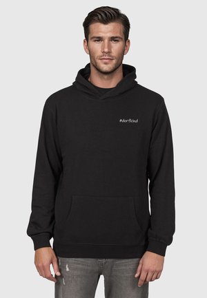 Ung mann iført svart hettegenser med frontlomme og teksten "#dorfkind" på brystet, kombinert med grå slitte jeans.