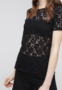 Top in pizzo nero con maniche corte, caratterizzato da motivi floreali e un design testurizzato, sovrapposto a una base nera uniforme.