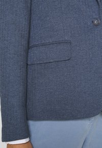 Blazer en bleu marine à motif en chevrons, avec un bouton unique, un col cranté et une poche plaquée à l'avant, fabriqué en tissu texturé.