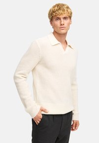 Maglione bianco a maglia con una trama testurizzata, scollatura a V e colletto ripiegato. Maniche lunghe con polsini larghi, abbinato a pantaloni grigio scuro.