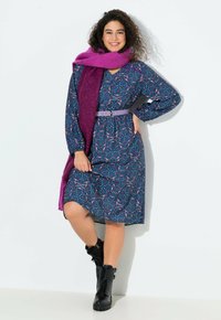 Frau in einem gemusterten lila Kleid mit einem fliederfarbenen Gürtel, einem dunkelroten Schal und schwarzen Kampfstiefeln, lächelt mit einer Hand auf der Hüfte.