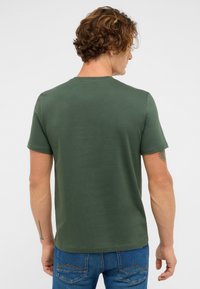 Homme avec des cheveux bouclés bruns portant un t-shirt vert uni à manches courtes et un jean bleu, de dos contre un fond blanc.