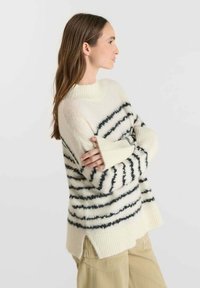 Maglione bianco accogliente con strisce orizzontali nere, tessuto morbido, polsini a coste e spacchi laterali. Indossato sopra pantaloni chiari.