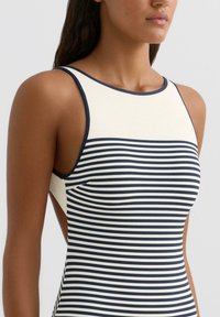 Gestreept badpak in marineblauw en crème. Heeft een hoge halslijn, mouwloos ontwerp en uitgesneden zijkanten. Materiaal lijkt van structuur te zijn.