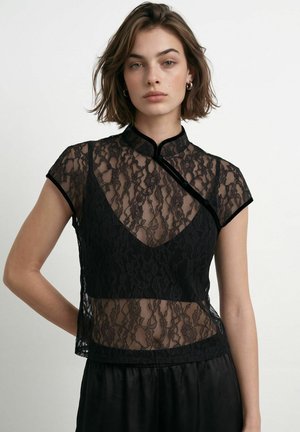 Jeune femme aux cheveux bruns courts portant un top noir en dentelle à col montant et manches courtes ainsi qu'une jupe noire sur un fond clair uni.