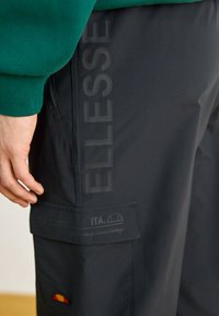 Zwarte cargobroek met een soepele textuur, voorzien van een opvallend "ELLESSE" logo aan de zijkant, een ritssluitingzak en een badge met merkinformatie.