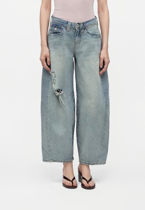 Person iført lyseblå jeans med brede ben og en revnet lap på venstre lår, lyst top og sorte hælsandaler, står med fronten fremad.