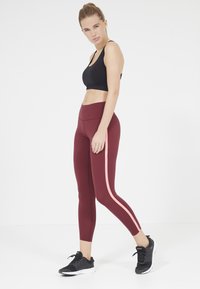 Leggings bordeaux con una striscia laterale rosa, realizzati in materiale elastico, abbinati a un reggiseno sportivo nero e a scarpe da ginnastica nere.